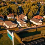 High-angle view of a residential neighborhood, with various-sized houses and properties of different shapes and sizes lining the streets. The scene is bathed in warm, golden afternoon sunlight, casting long shadows across the well-manicured lawns and neatly trimmed hedges. In the foreground, a "For Sale" sign stands prominently, drawing the viewer's attention to the varying property values represented by the diverse homes. The middle ground features a mix of modern and traditional architectural styles, while the background is framed by lush, mature trees swaying gently in a light breeze. The overall atmosphere conveys a sense of tranquility and the desirability of the neighborhood, underscoring the theme of "Side Property Prices".